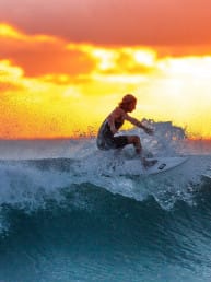 Man riding a surf board on the crest of a wave with sun setting behind